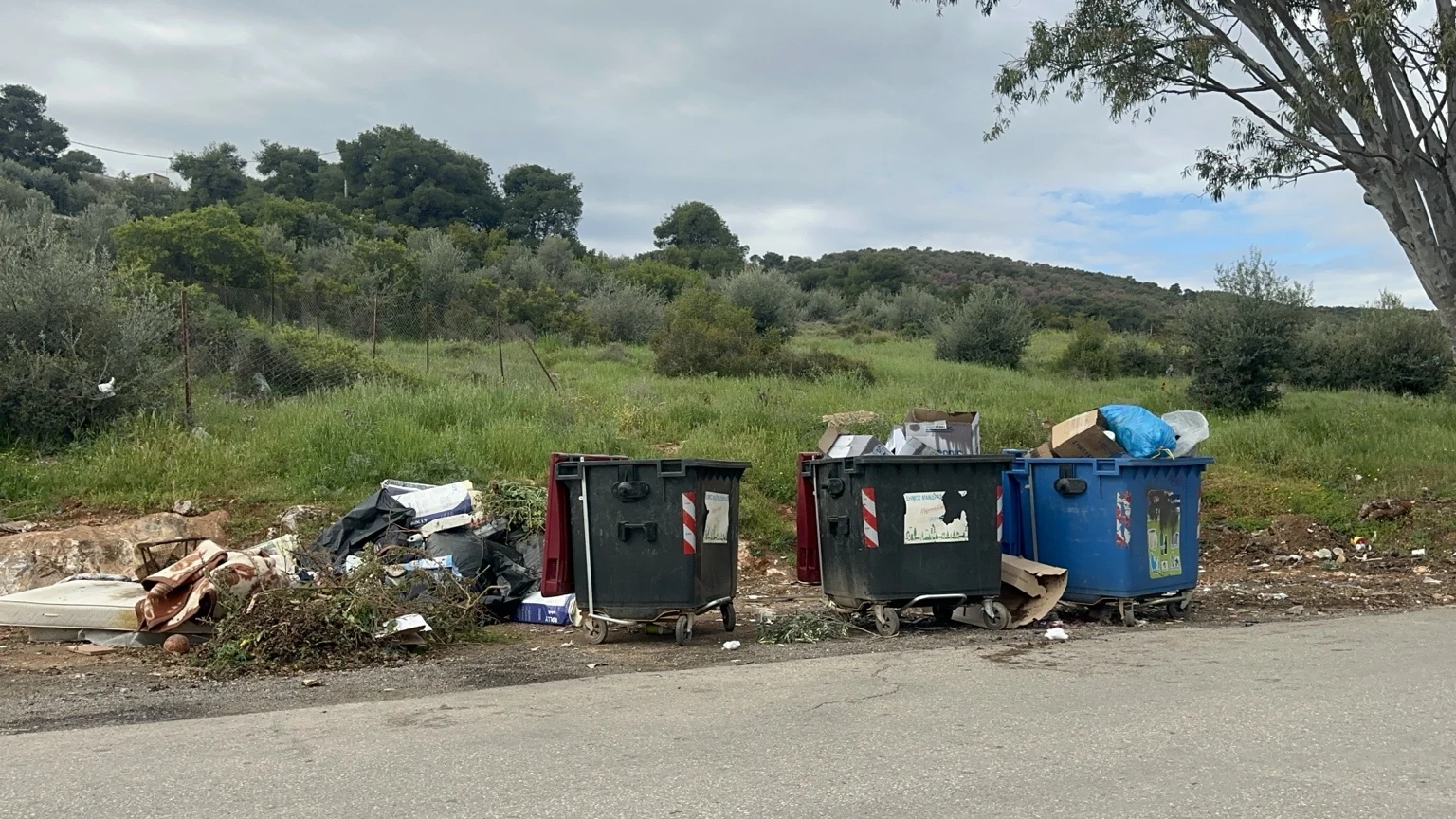Μπλε κάδος και δύο σκούροι κάδοι με υπερχείλιση υλικών και απορρίμματα στο έδαφος δίπλα σε βλάστηση.